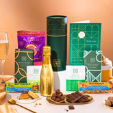 EDEN & CO. plant-based gift hamper with a mini Bottega Gold prosecco, green gift tube, “Occasion Themed” card, chocolate bars in Luscious Orange, White Chocolate Velvet, Signature Chocolate Blend and Salted Caramel, Booja-Booja Chocolate Wonders truffles, Buttermilk Peanut Nougat and Honeycomb Blast bars, chocolate truffles and honeycomb pieces on small plates, and a filled champagne glass, set against a warm terracotta background.