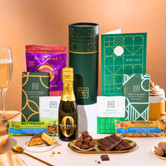 EDEN & CO. plant-based gift hamper with a mini Bottega Zero Black prosecco, green gift tube, “Occasion Themed” card, chocolate bars in Luscious Orange, White Chocolate Velvet, Signature Chocolate Blend and Salted Caramel, Booja-Booja Chocolate Wonders truffles, Buttermilk Peanut Nougat and Honeycomb Blast bars, chocolate truffles and honeycomb pieces on small plates, and a filled champagne glass, set against a warm terracotta background.