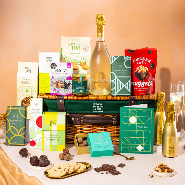 EDEN & CO. Luxury Wicker Hamper filled with a full-size Bottega Gold prosecco, an assortment of chocolate bars, Booja-Booja Chocolate Wonders truffles and Buttermilk snack bars, presented in a dark green trimmed wicker basket with an “Occasion Themed” card, styled against a warm terracotta background.