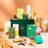EDEN & CO. Food & Drink Hamper with a full-size Bottega Gold prosecco, dark green gift cylinder, an “Occasion Themed” card, an assortment of chocolate bars, Booja-Booja Chocolate Wonders truffles, Buttermilk snack bars, a scented candle and Aery hand cream, styled with gold silk against a warm terracotta background.