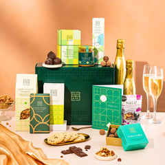 EDEN & CO. Food & Drink Hamper with a full-size Bottega Gold prosecco, dark green gift cylinder, an “Occasion Themed” card, an assortment of chocolate bars, Booja-Booja Chocolate Wonders truffles, Buttermilk snack bars, a scented candle and Aery hand cream, styled with gold silk against a warm terracotta background.