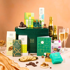 EDEN & CO. Food & Drink Hamper with a full-size Bottega Zero Black prosecco (0% alcohol-free), dark green gift cylinder, an “Occasion Themed” card, an assortment of chocolate bars, Booja-Booja Chocolate Wonders truffles, Buttermilk snack bars, a scented candle and Aery hand cream, styled with gold silk against a warm terracotta background.