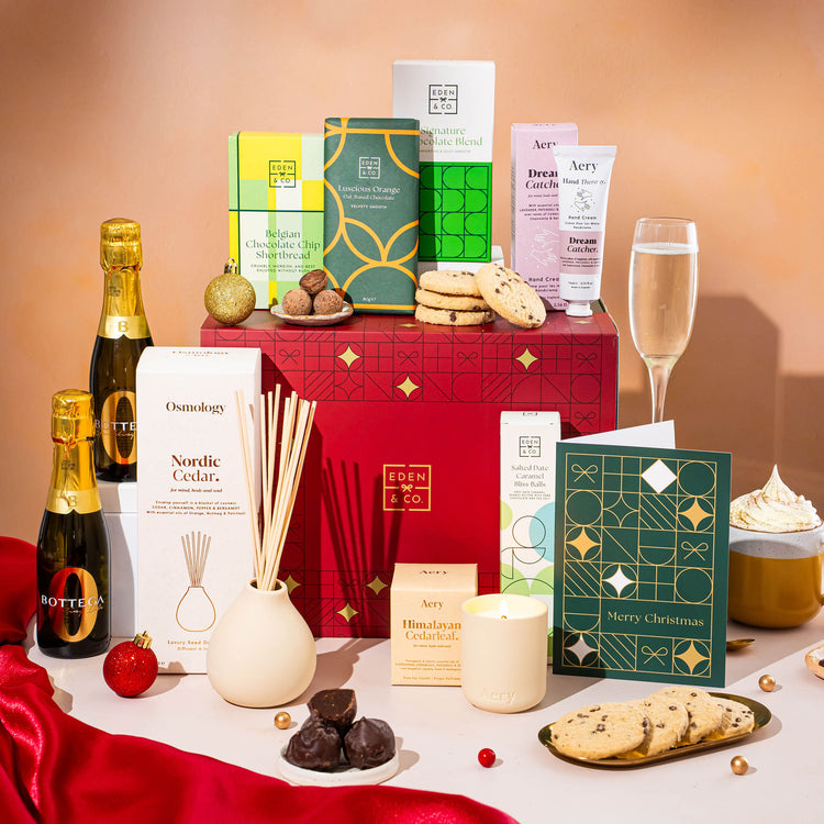 EDEN & CO. Pamper Hamper gift set with a mini Bottega Zero Black prosecco (0% alcohol-free), dark green gift cylinder, an “Occasion Themed” card, chocolate bars and truffles, a scented candle and Aery hand cream, styled with gold silk against a warm terracotta background.