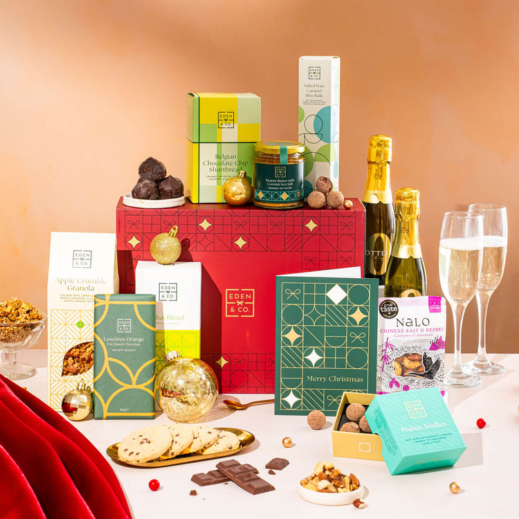 EDEN & CO. Food & Drink Hamper with a full-size Bottega Zero Black prosecco (0% alcohol-free), dark green gift cylinder, an “Occasion Themed” card, an assortment of chocolate bars, Booja-Booja Chocolate Wonders truffles, Buttermilk snack bars, a scented candle and Aery hand cream, styled with gold silk against a warm terracotta background.