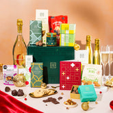 EDEN & CO. Grand Hamper gift box with a full-size Bottega Gold prosecco, dark green gift cylinder, an “Occasion Themed” card, an assortment of chocolate bars, Booja-Booja Chocolate Wonders truffles and Buttermilk snack bars, styled with gold silk against a warm terracotta background.