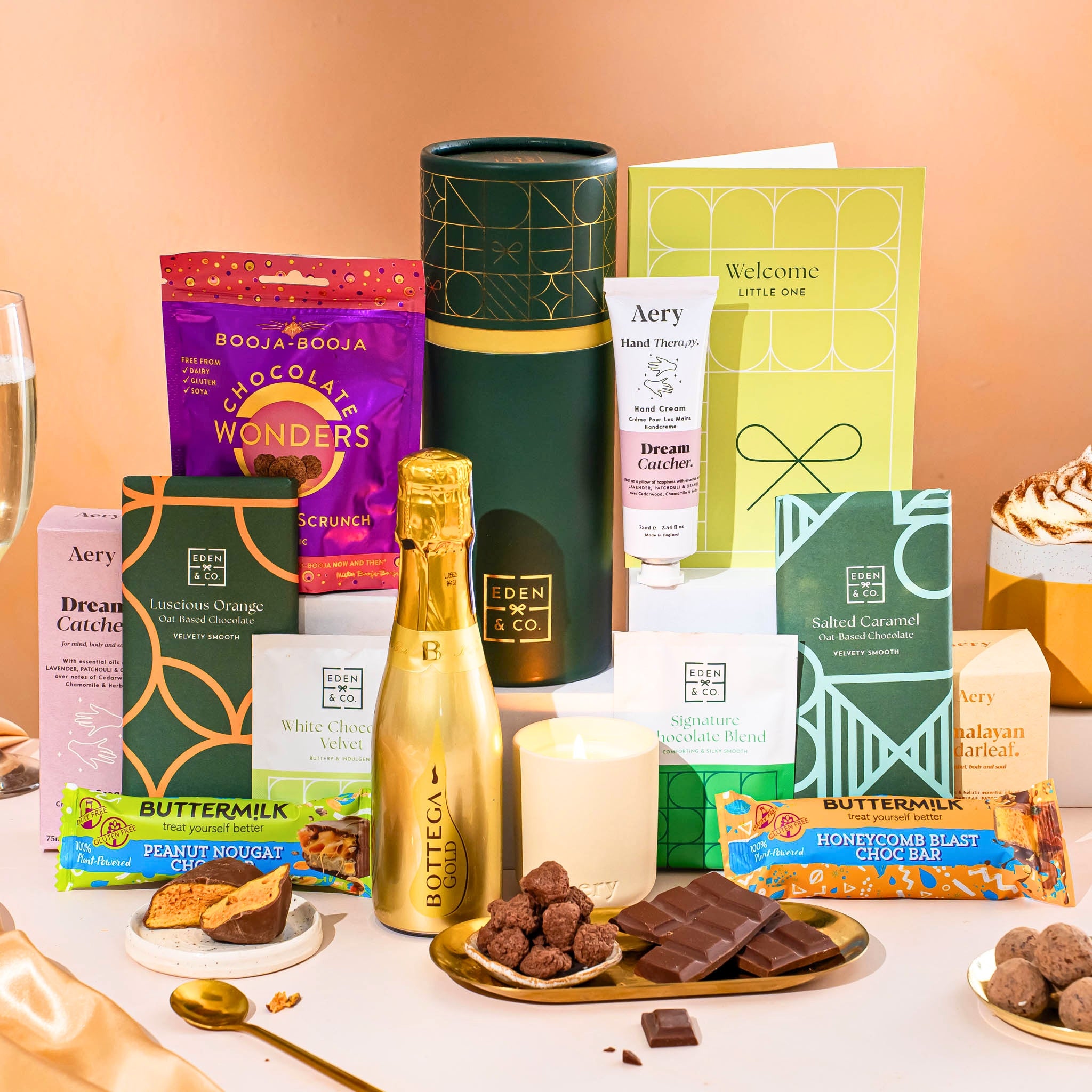 EDEN & CO. plant-based gift hamper with a mini Bottega Gold prosecco, green gift tube, “Occasion Themed” card, chocolate bars in Luscious Orange, White Chocolate Velvet, Signature Chocolate Blend and Salted Caramel, Booja-Booja Chocolate Wonders truffles, Buttermilk Peanut Nougat and Honeycomb Blast bars, chocolate truffles and honeycomb pieces on small plates, and a filled champagne glass, luxury candle and hand cream set against a warm terracotta background.