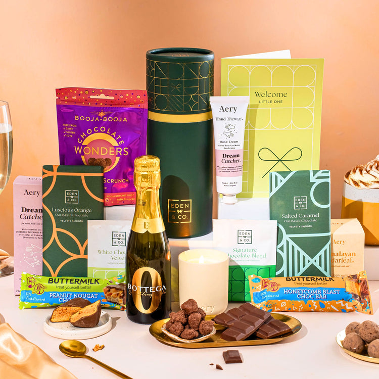 EDEN & CO. plant-based gift hamper with a mini Bottega Zero Black prosecco, green gift tube, “Occasion Themed” card, chocolate bars in Luscious Orange, White Chocolate Velvet, Signature Chocolate Blend and Salted Caramel, Booja-Booja Chocolate Wonders truffles, Buttermilk Peanut Nougat and Honeycomb Blast bars, chocolate truffles and honeycomb pieces on small plates, and a filled champagne glass,  luxury candle and hand cream set against a warm terracotta background.
