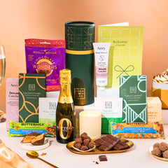 EDEN & CO. plant-based gift hamper with a mini Bottega Zero Black prosecco, green gift tube, “Occasion Themed” card, chocolate bars in Luscious Orange, White Chocolate Velvet, Signature Chocolate Blend and Salted Caramel, Booja-Booja Chocolate Wonders truffles, Buttermilk Peanut Nougat and Honeycomb Blast bars, chocolate truffles and honeycomb pieces on small plates, and a filled champagne glass,  luxury candle and hand cream set against a warm terracotta background.