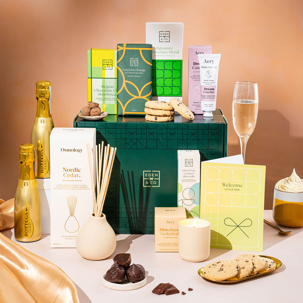EDEN & CO. Pamper Hamper gift set with a mini Bottega Gold prosecco, dark green gift cylinder, an “Occasion Themed” card, chocolate bars and truffles, a scented candle and Aery hand cream, styled with gold silk against a warm terracotta background.