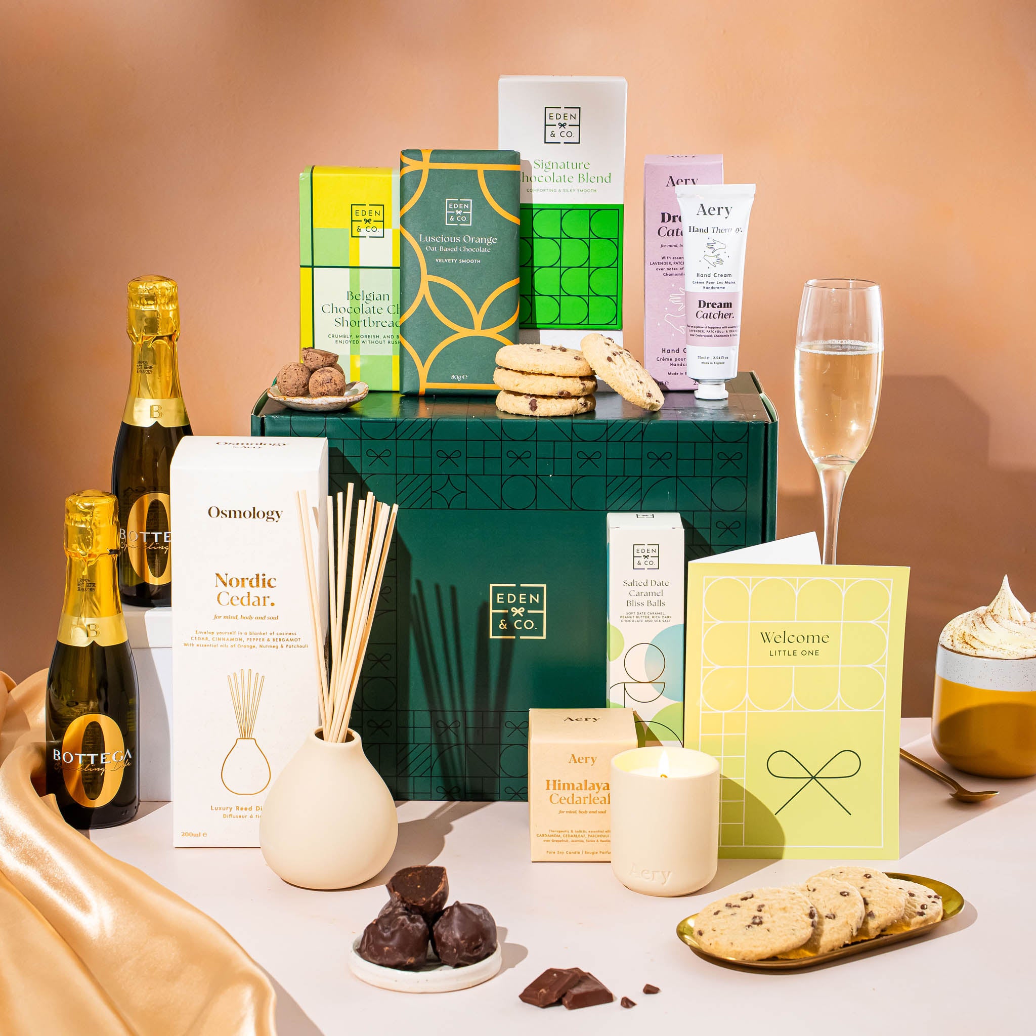 EDEN & CO. Pamper Hamper gift set with a mini Bottega Zero Black prosecco (0% alcohol-free), dark green gift cylinder, an “Occasion Themed” card, chocolate bars and truffles, a scented candle and Aery hand cream, styled with gold silk against a warm terracotta background.