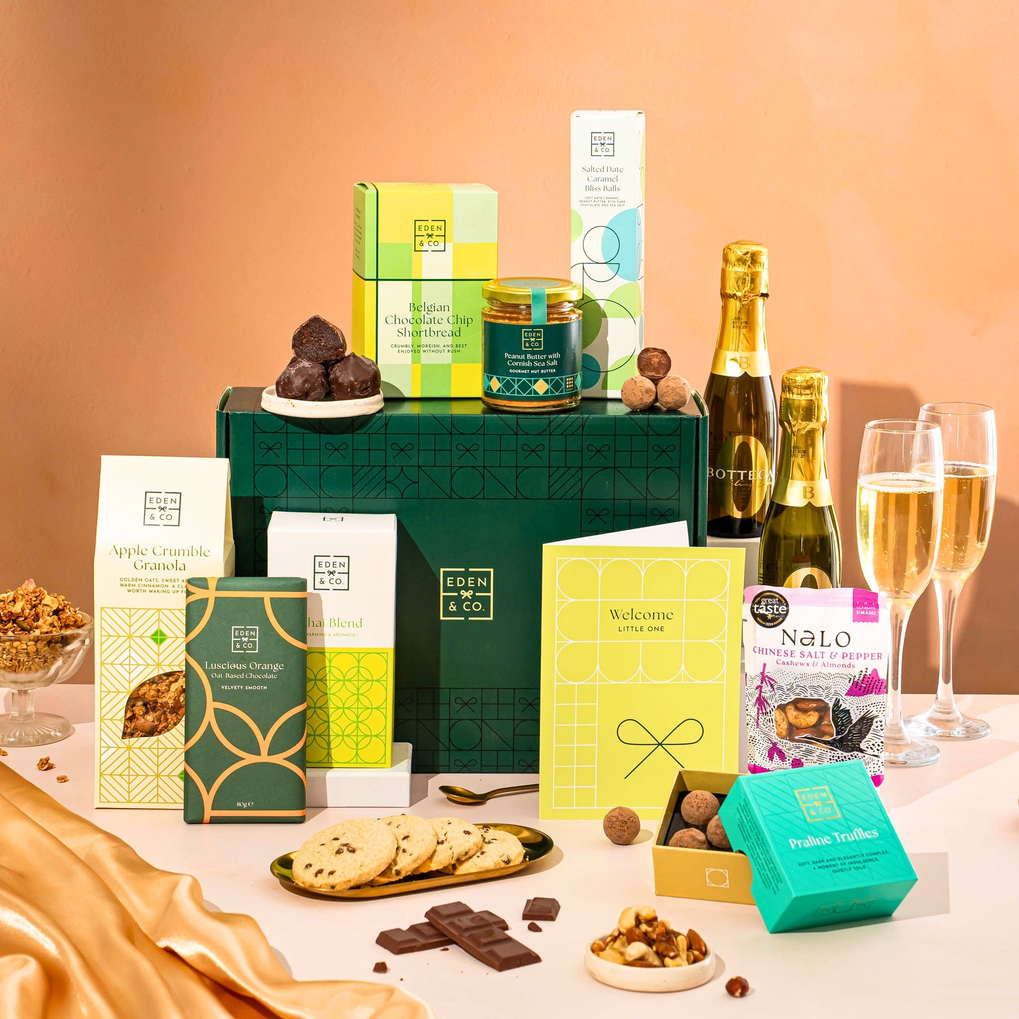 EDEN & CO. Food & Drink Hamper with a full-size Bottega Zero Black prosecco (0% alcohol-free), dark green gift cylinder, an “Occasion Themed” card, an assortment of chocolate bars, Booja-Booja Chocolate Wonders truffles, Buttermilk snack bars, a scented candle and Aery hand cream, styled with gold silk against a warm terracotta background.