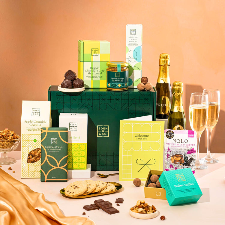 EDEN & CO. Food & Drink Hamper with a full-size Bottega Zero Black prosecco (0% alcohol-free), dark green gift cylinder, an “Occasion Themed” card, an assortment of chocolate bars, Booja-Booja Chocolate Wonders truffles, Buttermilk snack bars, a scented candle and Aery hand cream, styled with gold silk against a warm terracotta background.