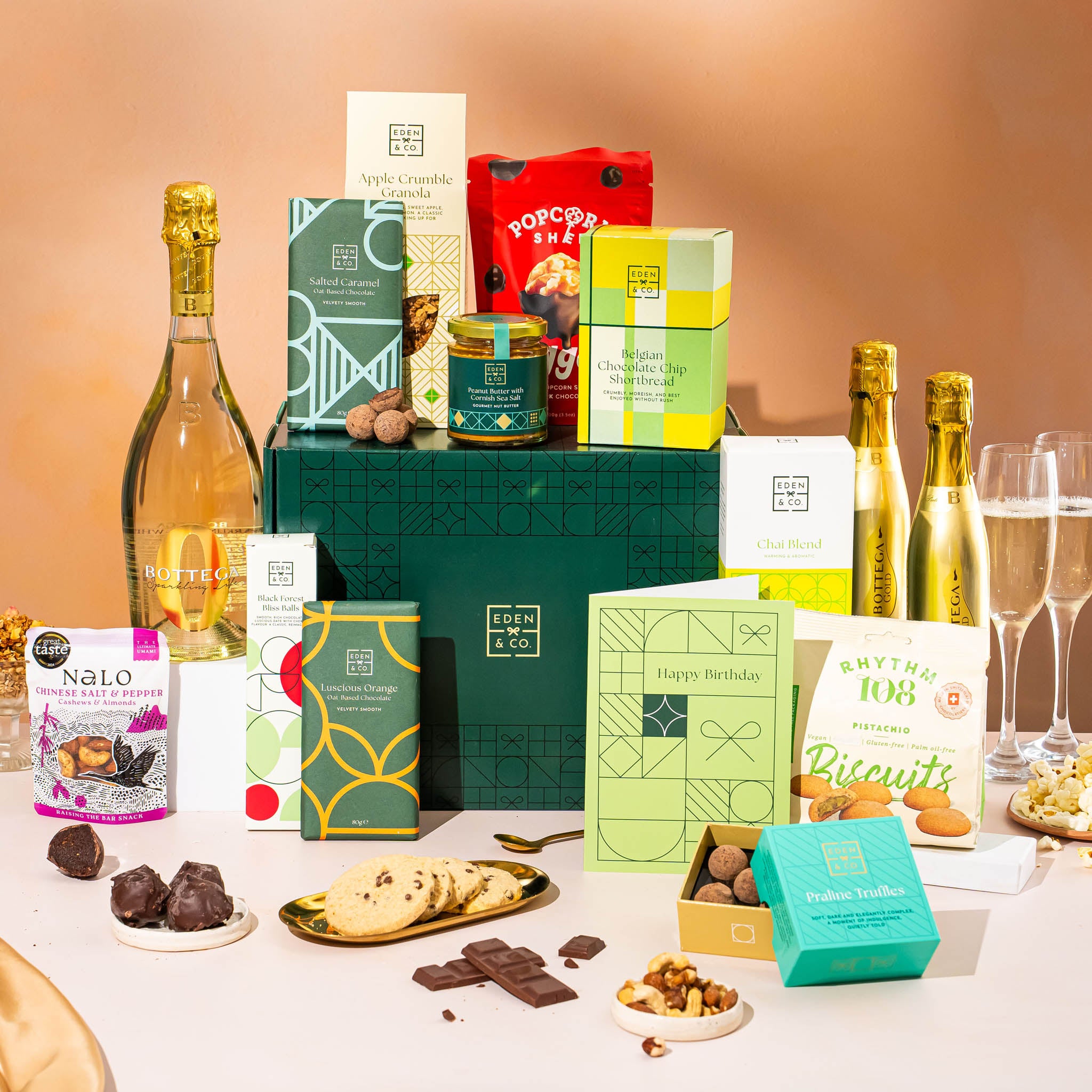 EDEN & CO. Grand Hamper gift box with a full-size Bottega Gold prosecco, dark green gift cylinder, an “Occasion Themed” card, an assortment of chocolate bars, Booja-Booja Chocolate Wonders truffles and Buttermilk snack bars, styled with gold silk against a warm terracotta background.