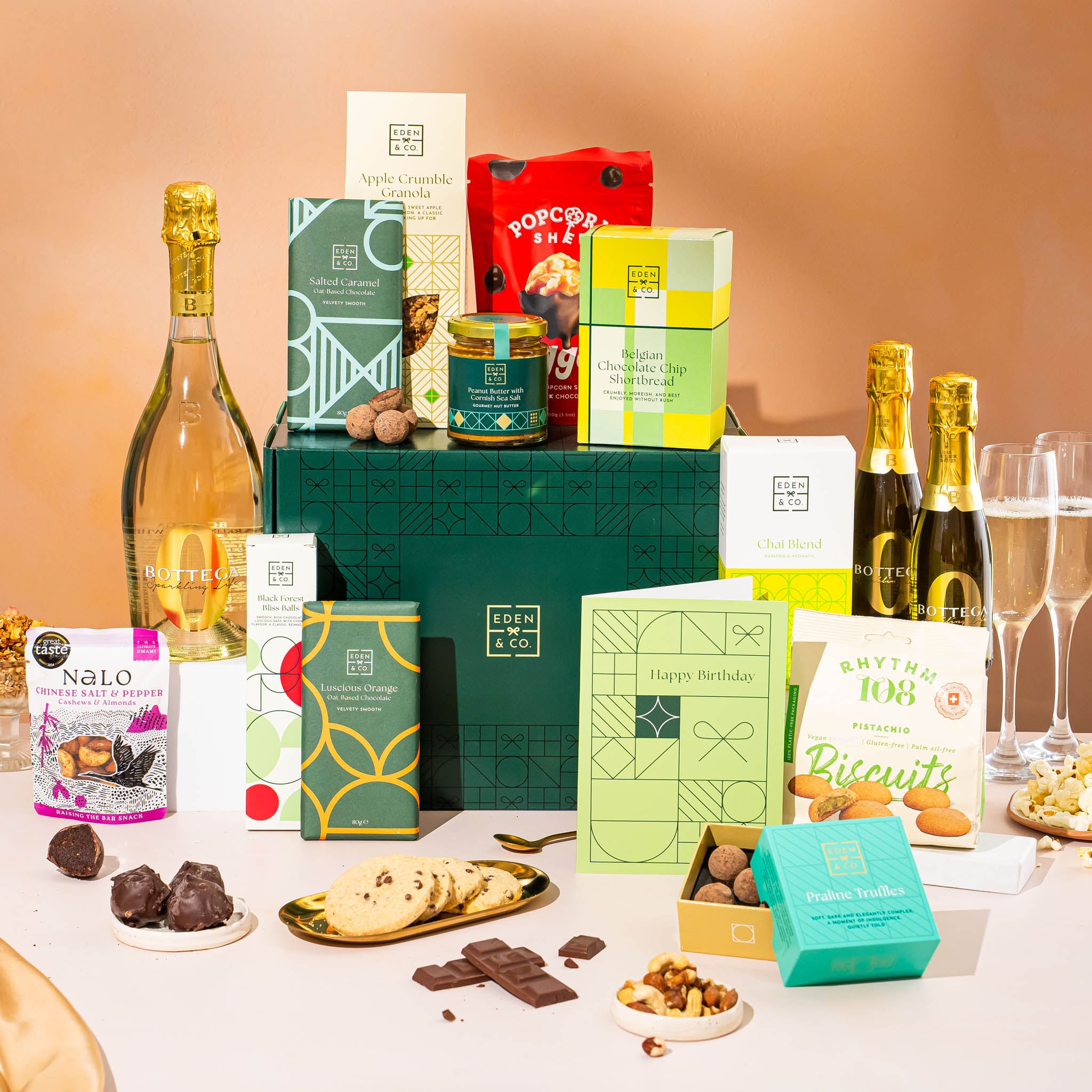 EDEN & CO. Grand Hamper gift box with a full-size Bottega Zero Black prosecco (0% alcohol-free), dark green gift cylinder, an “Occasion Themed” card, an assortment of chocolate bars, Booja-Booja Chocolate Wonders truffles and Buttermilk snack bars, styled with gold silk against a warm terracotta background.