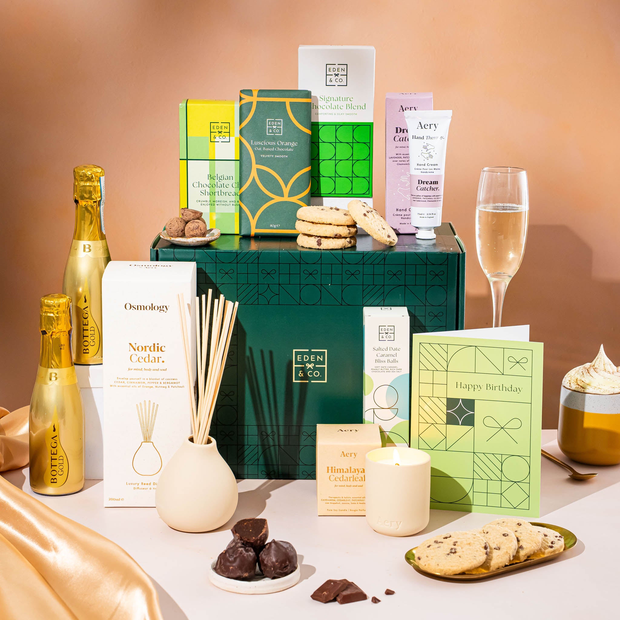 EDEN & CO. Pamper Hamper gift set with a mini Bottega Gold prosecco, dark green gift cylinder, an “Occasion Themed” card, chocolate bars and truffles, a scented candle and Aery hand cream, styled with gold silk against a warm terracotta background.
