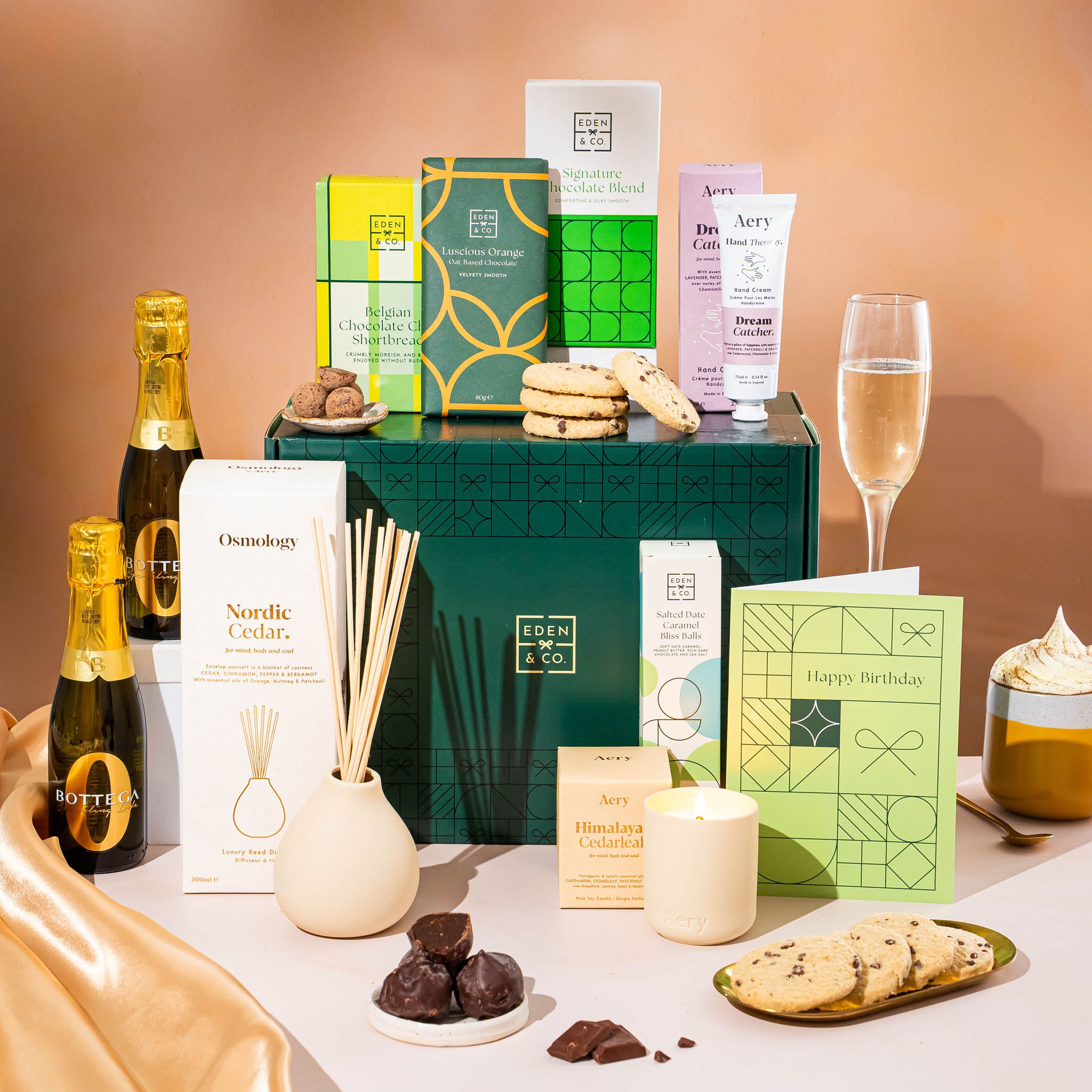 EDEN & CO. Pamper Hamper gift set with a mini Bottega Zero Black prosecco (0% alcohol-free), dark green gift cylinder, an “Occasion Themed” card, chocolate bars and truffles, a scented candle and Aery hand cream, styled with gold silk against a warm terracotta background.
