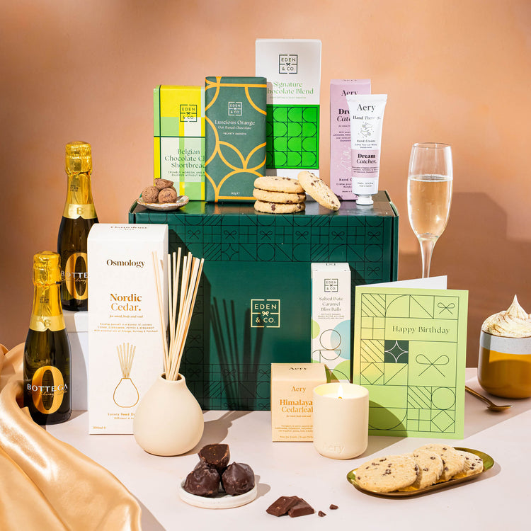 EDEN & CO. Pamper Hamper gift set with a mini Bottega Zero Black prosecco (0% alcohol-free), dark green gift cylinder, an “Occasion Themed” card, chocolate bars and truffles, a scented candle and Aery hand cream, styled with gold silk against a warm terracotta background.