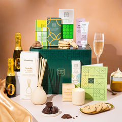 EDEN & CO. Pamper Hamper gift set with a mini Bottega Zero Black prosecco (0% alcohol-free), dark green gift cylinder, an “Occasion Themed” card, chocolate bars and truffles, a scented candle and Aery hand cream, styled with gold silk against a warm terracotta background.