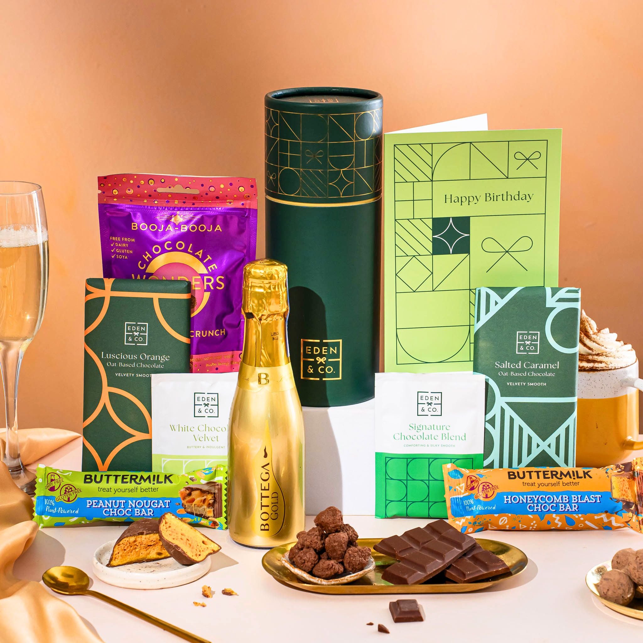 EDEN & CO. plant-based gift hamper with a mini Bottega Gold prosecco, green gift tube, “Occasion Themed” card, chocolate bars in Luscious Orange, White Chocolate Velvet, Signature Chocolate Blend and Salted Caramel, Booja-Booja Chocolate Wonders truffles, Buttermilk Peanut Nougat and Honeycomb Blast bars, chocolate truffles and honeycomb pieces on small plates, and a filled champagne glass, set against a warm terracotta background.