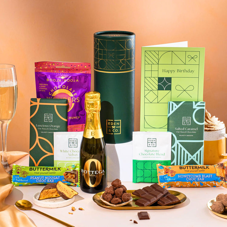 EDEN & CO. plant-based gift hamper with a mini Bottega Zero Black prosecco, green gift tube, “Occasion Themed” card, chocolate bars in Luscious Orange, White Chocolate Velvet, Signature Chocolate Blend and Salted Caramel, Booja-Booja Chocolate Wonders truffles, Buttermilk Peanut Nougat and Honeycomb Blast bars, chocolate truffles and honeycomb pieces on small plates, and a filled champagne glass, set against a warm terracotta background.