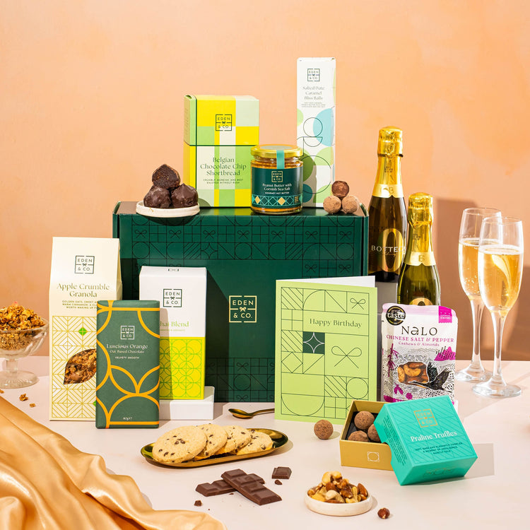 EDEN & CO. Food & Drink Hamper with a full-size Bottega Zero Black prosecco (0% alcohol-free), dark green gift cylinder, an “Occasion Themed” card, an assortment of chocolate bars, Booja-Booja Chocolate Wonders truffles, Buttermilk snack bars, a scented candle and Aery hand cream, styled with gold silk against a warm terracotta background.