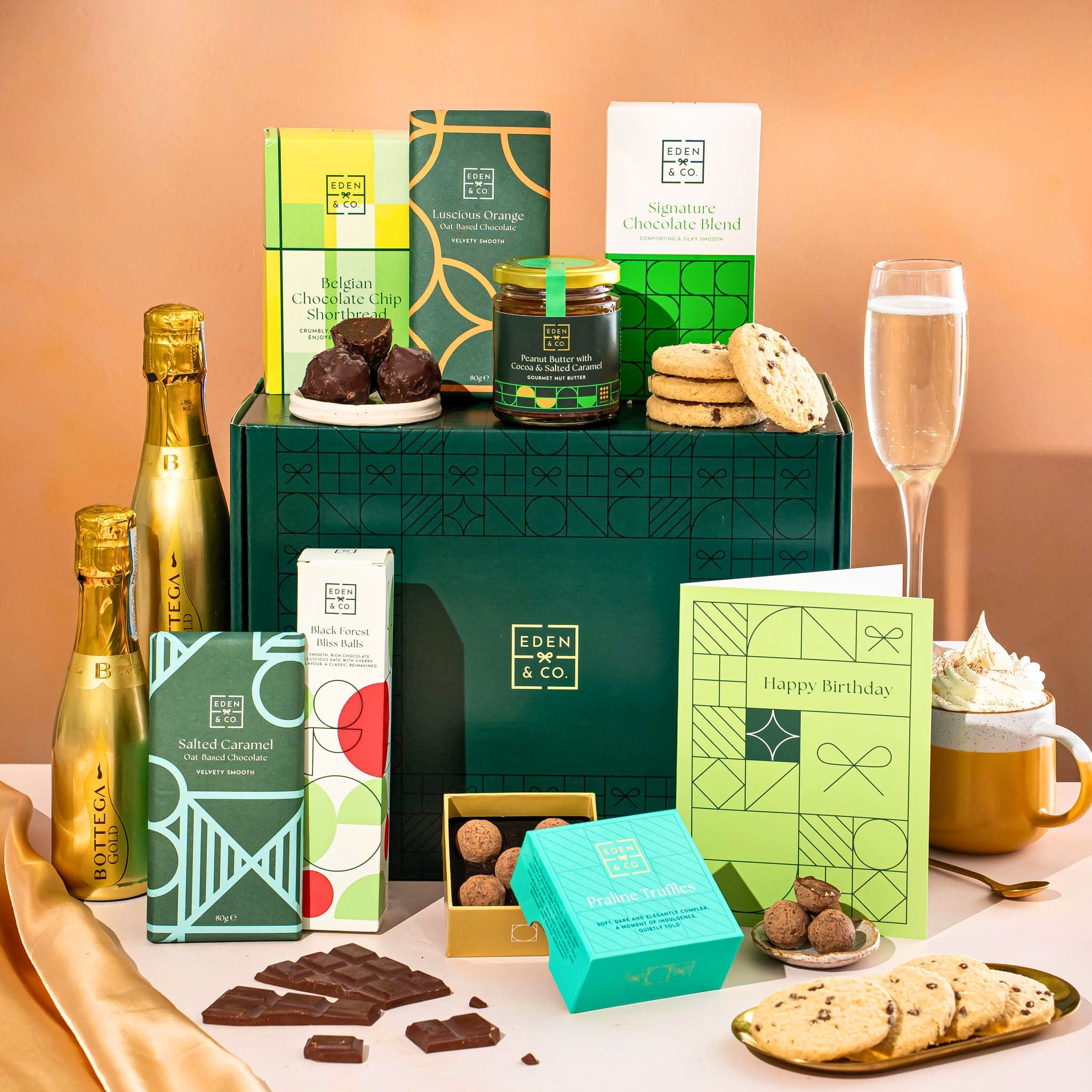 EDEN & CO. Chocolate Hamper letterbox gift with a mini Bottega Gold prosecco, green gift tube, an “Occasion Themed” card, an assortment of chocolate bars, Booja-Booja Chocolate Wonders truffles and chocolate truffles on a small plate, styled against a warm terracotta background.