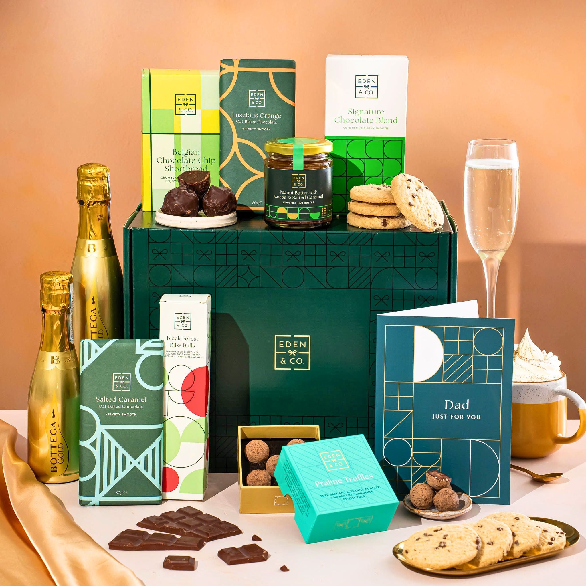 EDEN & CO. Chocolate Hamper letterbox gift with a mini Bottega Gold prosecco, green gift tube, an “Occasion Themed” card, an assortment of chocolate bars, Booja-Booja Chocolate Wonders truffles and chocolate truffles on a small plate, styled against a warm terracotta background.