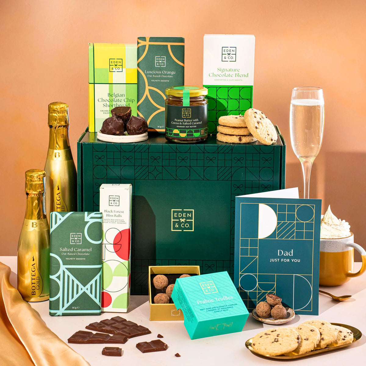 EDEN & CO. Chocolate Hamper letterbox gift with a mini Bottega Gold prosecco, green gift tube, an “Occasion Themed” card, an assortment of chocolate bars, Booja-Booja Chocolate Wonders truffles and chocolate truffles on a small plate, styled against a warm terracotta background.