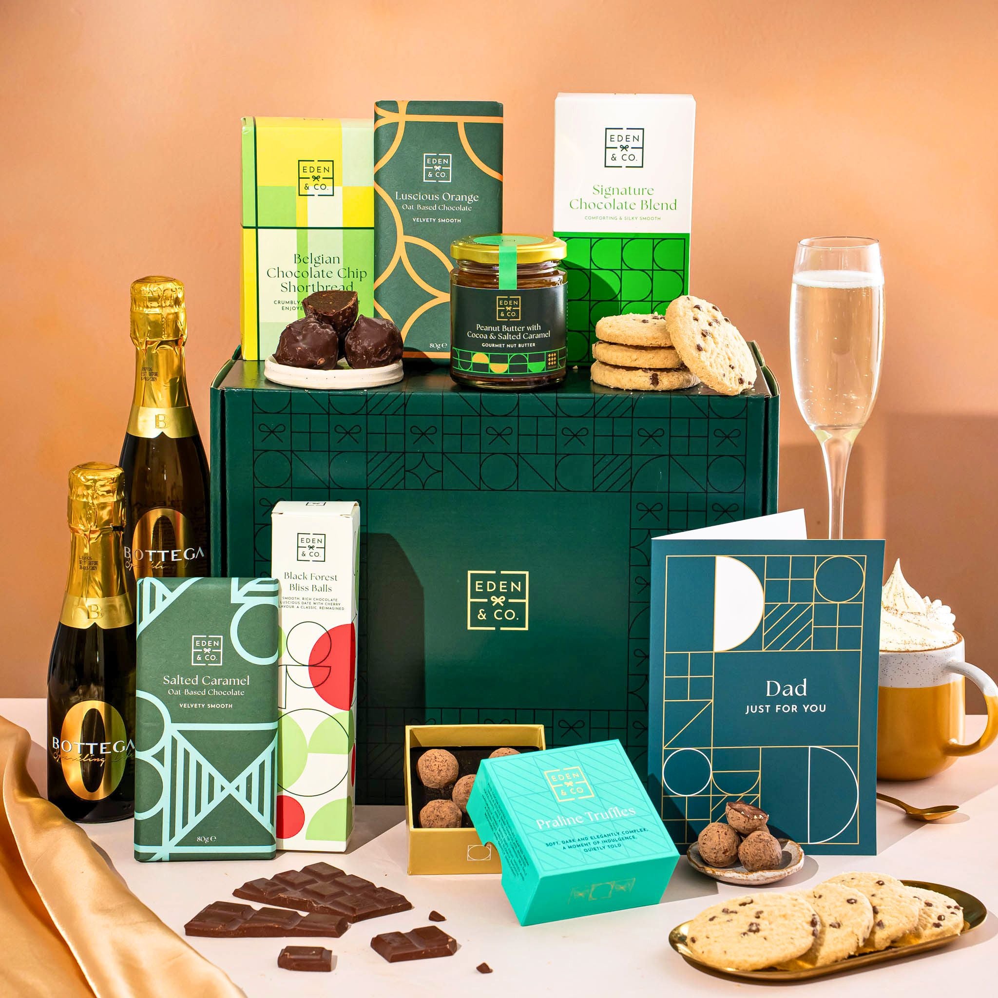 EDEN & CO. Chocolate Hamper letterbox gift with a mini Bottega Zero Black prosecco (0% alcohol-free), green gift tube, an “Occasion Themed” card, an assortment of chocolate bars, Booja-Booja Chocolate Wonders truffles and chocolate truffles on a small plate, styled against a warm terracotta background.