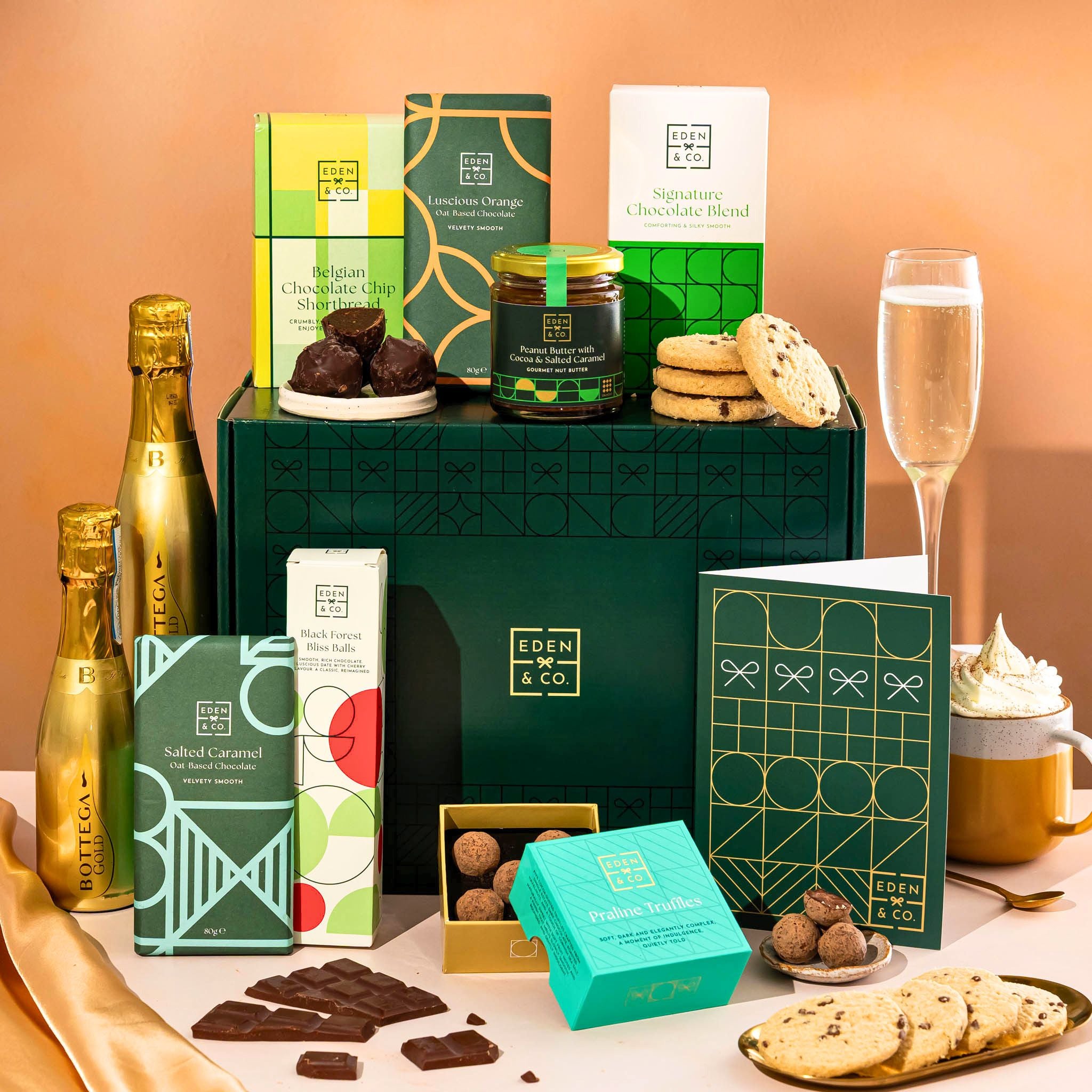 EDEN & CO. Chocolate Hamper letterbox gift with a mini Bottega Gold prosecco, green gift tube, an “Occasion Themed” card, an assortment of chocolate bars, Booja-Booja Chocolate Wonders truffles and chocolate truffles on a small plate, styled against a warm terracotta background.