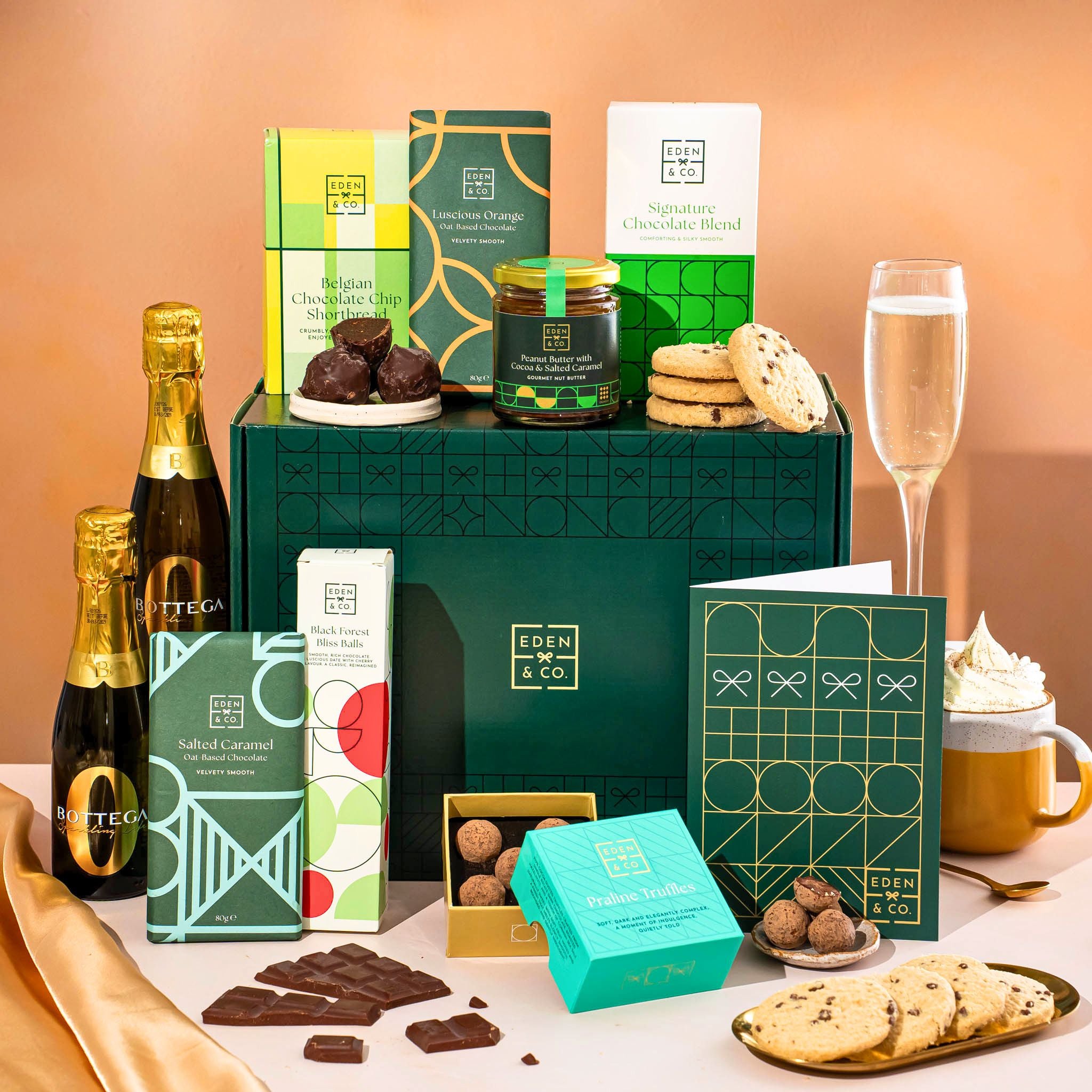 EDEN & CO. Chocolate Hamper letterbox gift with a mini Bottega Zero Black prosecco (0% alcohol-free), green gift tube, an “Occasion Themed” card, an assortment of chocolate bars, Booja-Booja Chocolate Wonders truffles and chocolate truffles on a small plate, styled against a warm terracotta background.