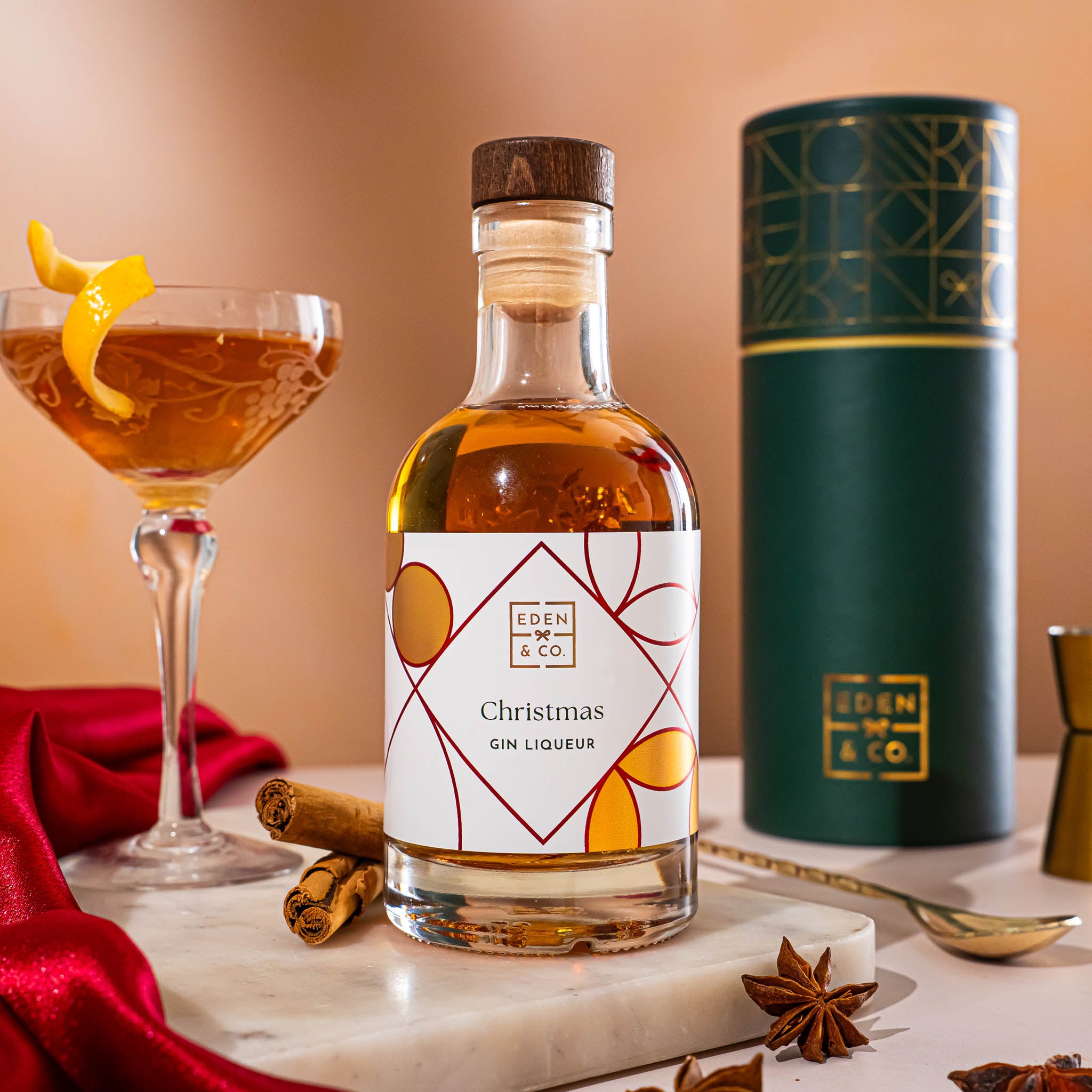 EDEN & CO. liqueur gift bottle with an “Occasion Themed” card, presented in a branded gift tube, styled with gold silk against a warm terracotta background.