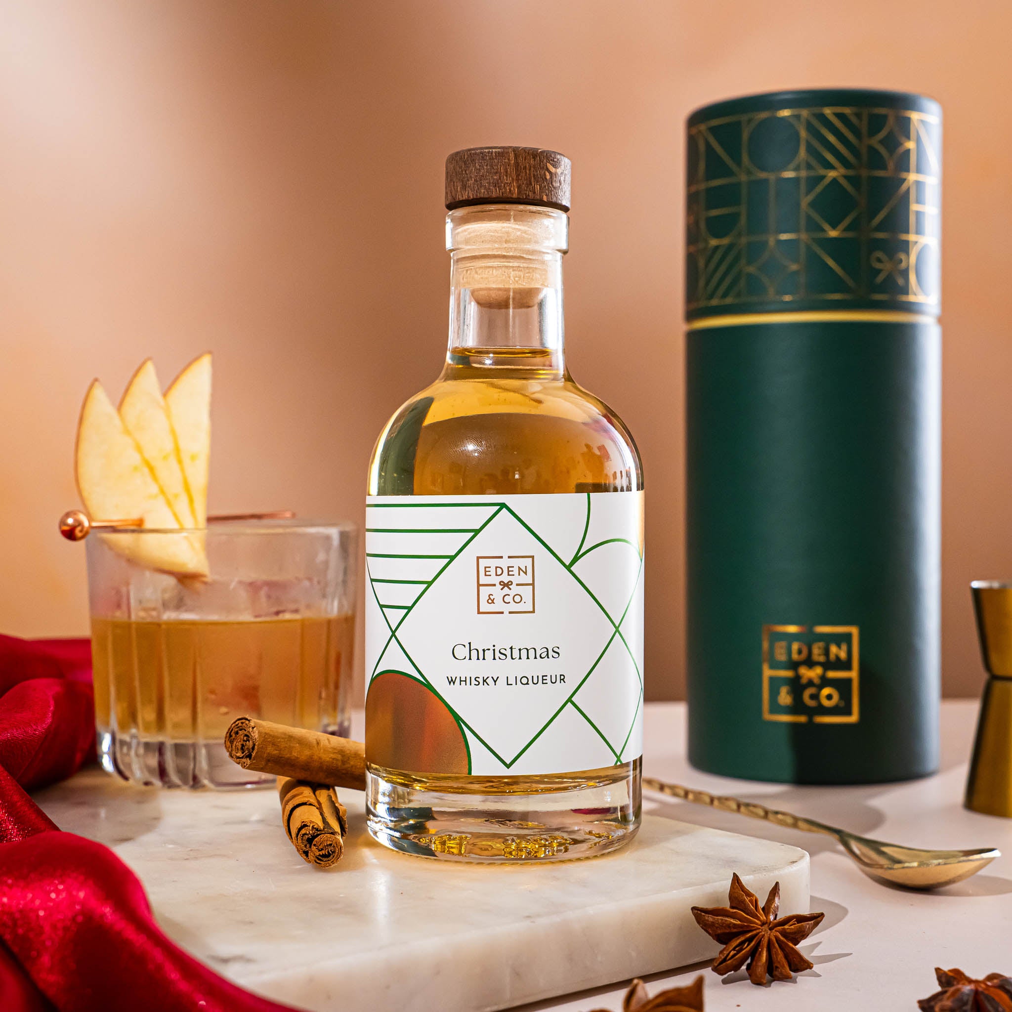 EDEN & CO. liqueur gift bottle with an “Occasion Themed” card, presented in a branded gift tube, styled with gold silk against a warm terracotta background.