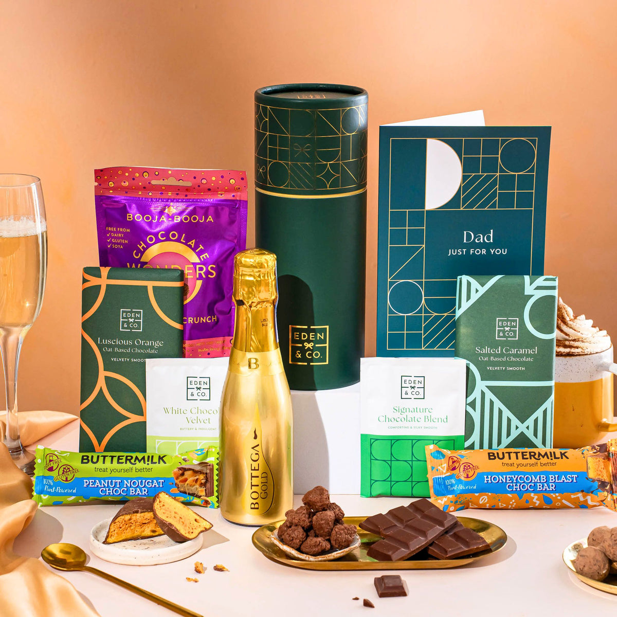 EDEN & CO. plant-based gift hamper with a mini Bottega Gold prosecco, green gift tube, “Occasion Themed” card, chocolate bars in Luscious Orange, White Chocolate Velvet, Signature Chocolate Blend and Salted Caramel, Booja-Booja Chocolate Wonders truffles, Buttermilk Peanut Nougat and Honeycomb Blast bars, chocolate truffles and honeycomb pieces on small plates, and a filled champagne glass, set against a warm terracotta background.