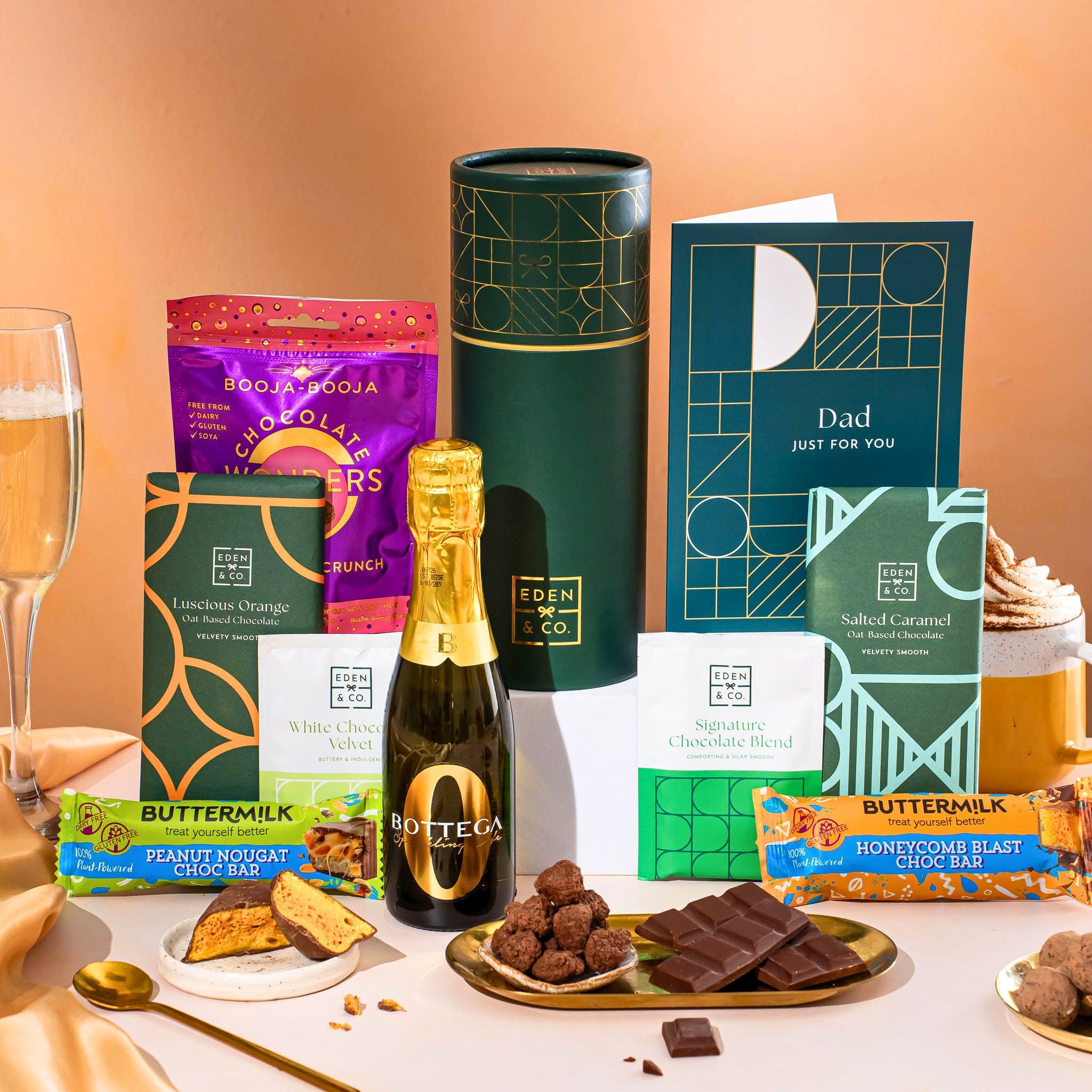 EDEN & CO. plant-based gift hamper with a mini Bottega Zero Black prosecco, green gift tube, “Occasion Themed” card, chocolate bars in Luscious Orange, White Chocolate Velvet, Signature Chocolate Blend and Salted Caramel, Booja-Booja Chocolate Wonders truffles, Buttermilk Peanut Nougat and Honeycomb Blast bars, chocolate truffles and honeycomb pieces on small plates, and a filled champagne glass, set against a warm terracotta background.