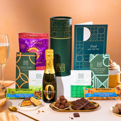EDEN & CO. plant-based gift hamper with a mini Bottega Zero Black prosecco, green gift tube, “Occasion Themed” card, chocolate bars in Luscious Orange, White Chocolate Velvet, Signature Chocolate Blend and Salted Caramel, Booja-Booja Chocolate Wonders truffles, Buttermilk Peanut Nougat and Honeycomb Blast bars, chocolate truffles and honeycomb pieces on small plates, and a filled champagne glass, set against a warm terracotta background.