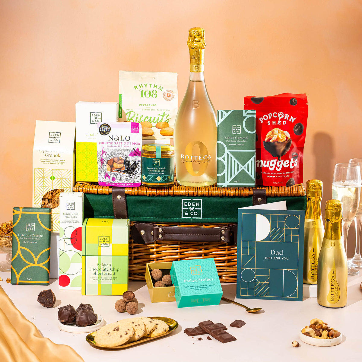 EDEN & CO. Luxury Wicker Hamper filled with a full-size Bottega Gold prosecco, an assortment of chocolate bars, Booja-Booja Chocolate Wonders truffles and Buttermilk snack bars, presented in a dark green trimmed wicker basket with an “Occasion Themed” card, styled against a warm terracotta background.
