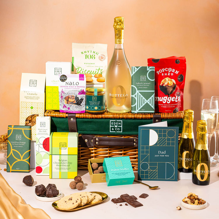 EDEN & CO. Luxury Wicker Hamper filled with a full-size Bottega Zero Black prosecco (0% alcohol-free), an assortment of chocolate bars, Booja-Booja Chocolate Wonders truffles and Buttermilk snack bars, presented in a dark green trimmed wicker basket with an “Occasion Themed” card, styled against a warm terracotta background.