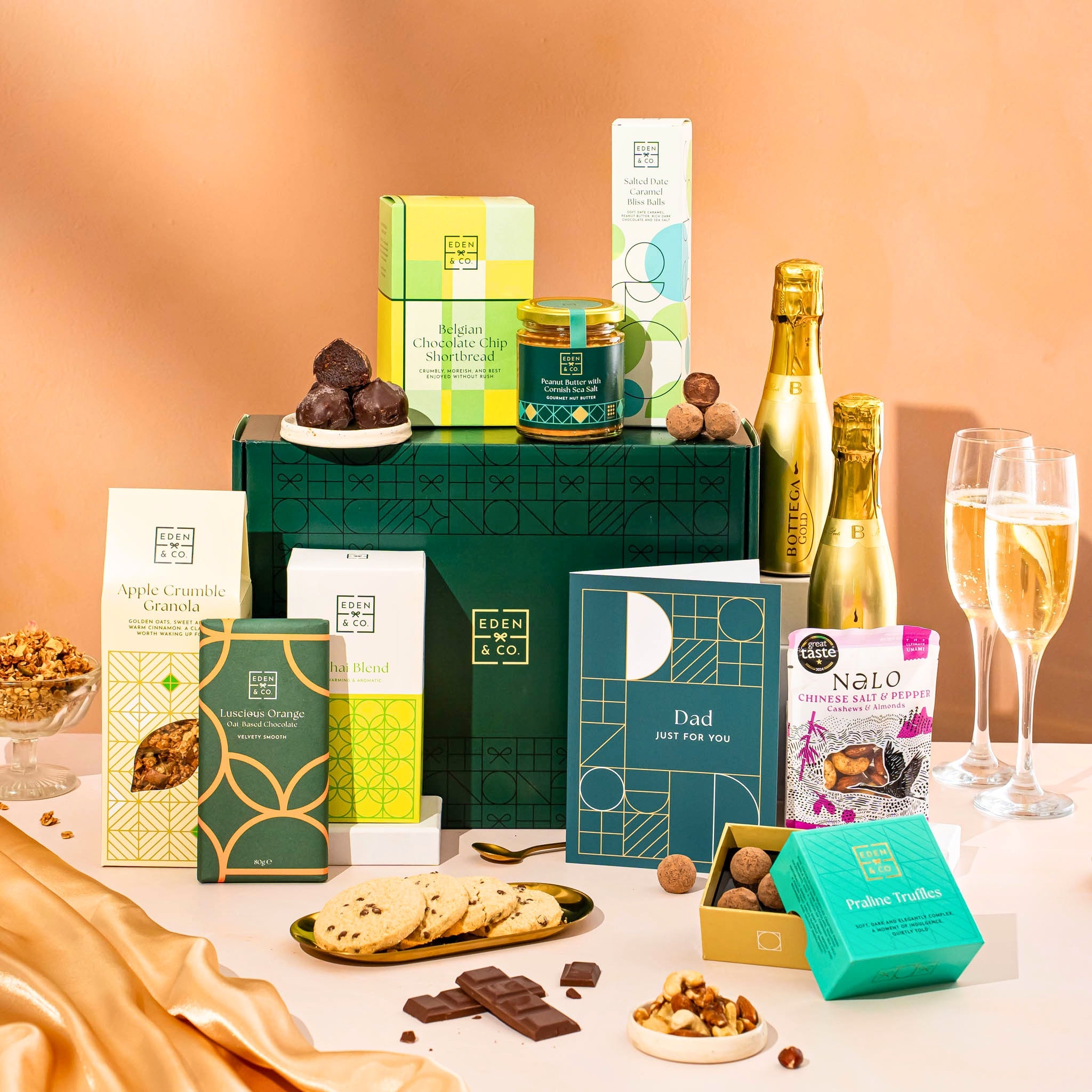 EDEN & CO. Food & Drink Hamper with a full-size Bottega Gold prosecco, dark green gift cylinder, an “Occasion Themed” card, an assortment of chocolate bars, Booja-Booja Chocolate Wonders truffles, Buttermilk snack bars, a scented candle and Aery hand cream, styled with gold silk against a warm terracotta background.
