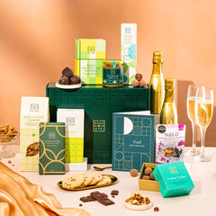 EDEN & CO. Food & Drink Hamper with a full-size Bottega Gold prosecco, dark green gift cylinder, an “Occasion Themed” card, an assortment of chocolate bars, Booja-Booja Chocolate Wonders truffles, Buttermilk snack bars, a scented candle and Aery hand cream, styled with gold silk against a warm terracotta background.