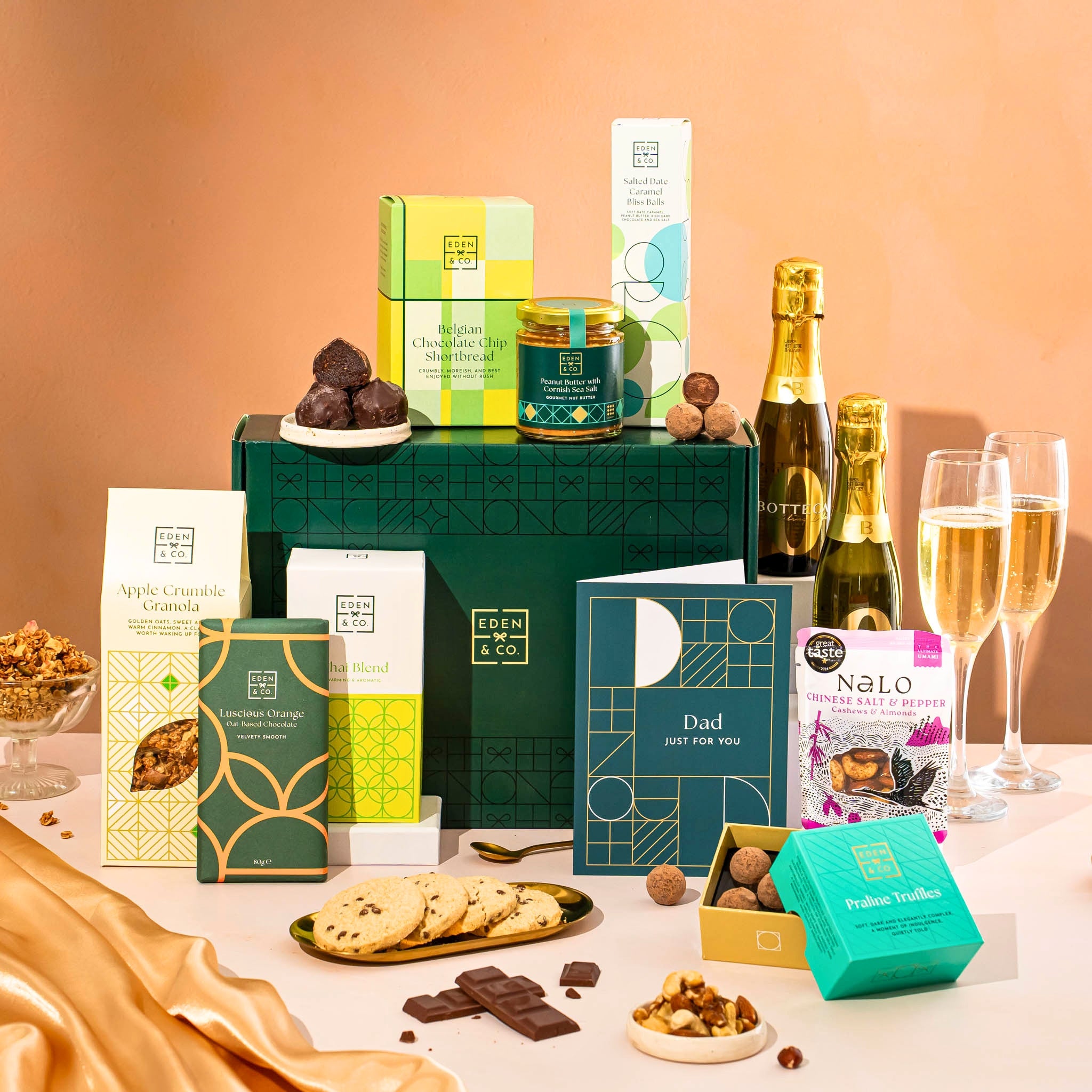 EDEN & CO. Food & Drink Hamper with a full-size Bottega Zero Black prosecco (0% alcohol-free), dark green gift cylinder, an “Occasion Themed” card, an assortment of chocolate bars, Booja-Booja Chocolate Wonders truffles, Buttermilk snack bars, a scented candle and Aery hand cream, styled with gold silk against a warm terracotta background.