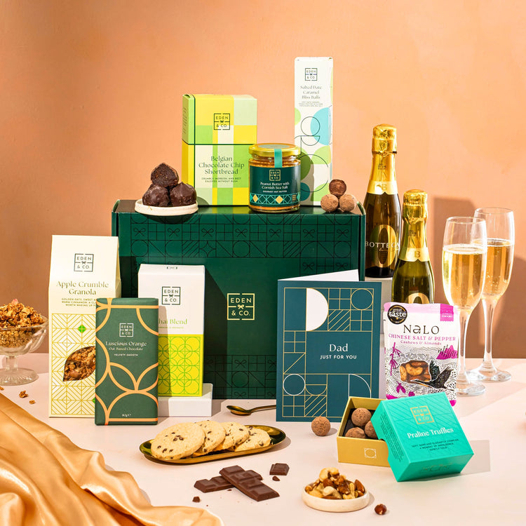 EDEN & CO. Food & Drink Hamper with a full-size Bottega Zero Black prosecco (0% alcohol-free), dark green gift cylinder, an “Occasion Themed” card, an assortment of chocolate bars, Booja-Booja Chocolate Wonders truffles, Buttermilk snack bars, a scented candle and Aery hand cream, styled with gold silk against a warm terracotta background.