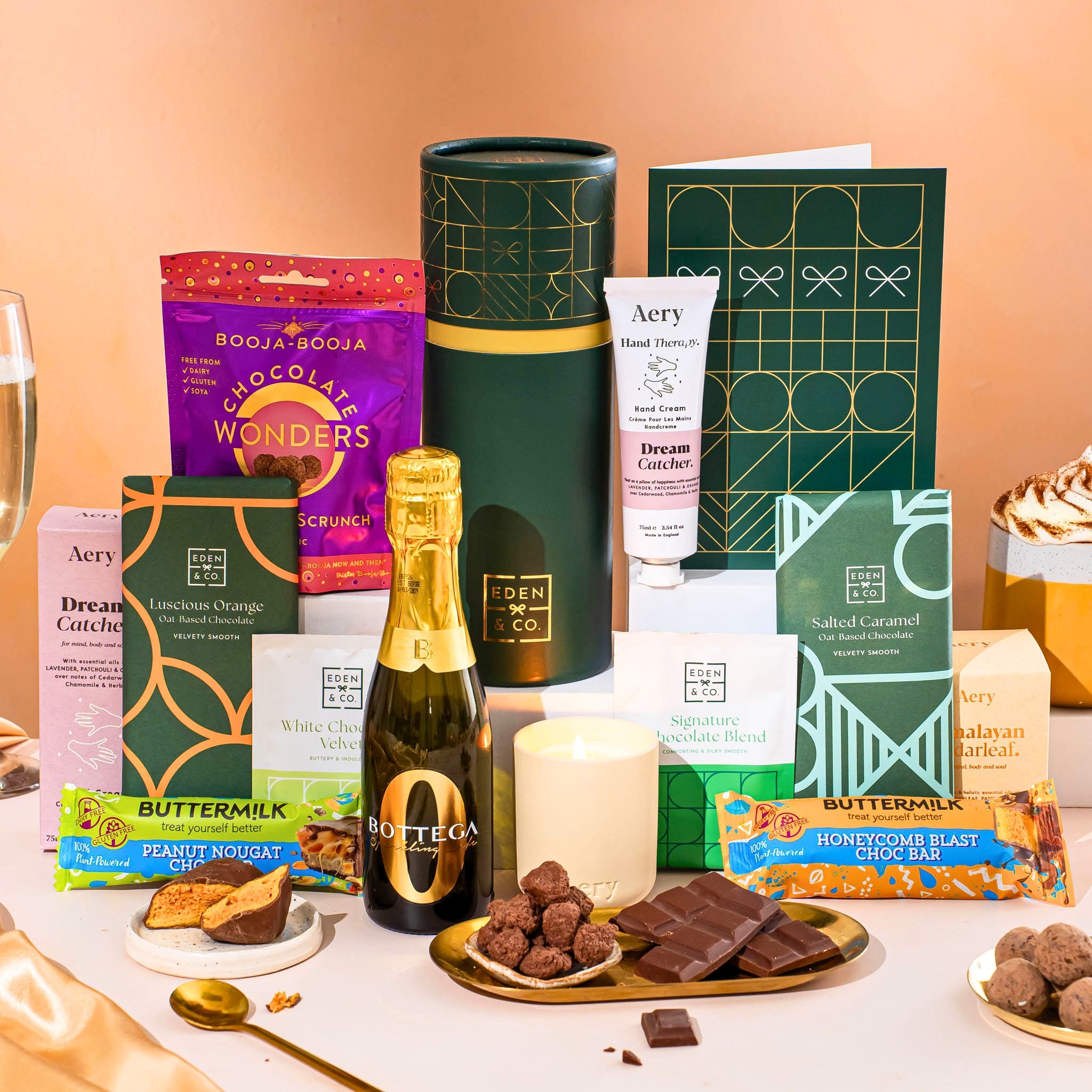 EDEN & CO. plant-based gift hamper with a mini Bottega Zero Black prosecco, green gift tube, “Occasion Themed” card, chocolate bars in Luscious Orange, White Chocolate Velvet, Signature Chocolate Blend and Salted Caramel, Booja-Booja Chocolate Wonders truffles, Buttermilk Peanut Nougat and Honeycomb Blast bars, chocolate truffles and honeycomb pieces on small plates, and a filled champagne glass,  luxury candle and hand cream set against a warm terracotta background.