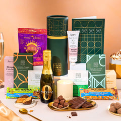 EDEN & CO. plant-based gift hamper with a mini Bottega Zero Black prosecco, green gift tube, “Occasion Themed” card, chocolate bars in Luscious Orange, White Chocolate Velvet, Signature Chocolate Blend and Salted Caramel, Booja-Booja Chocolate Wonders truffles, Buttermilk Peanut Nougat and Honeycomb Blast bars, chocolate truffles and honeycomb pieces on small plates, and a filled champagne glass,  luxury candle and hand cream set against a warm terracotta background.