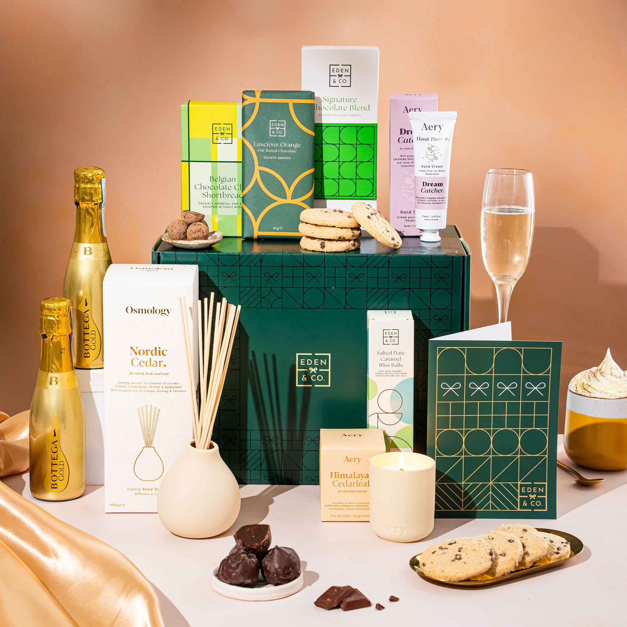 EDEN & CO. Pamper Hamper gift set with a mini Bottega Gold prosecco, dark green gift cylinder, an “Occasion Themed” card, chocolate bars and truffles, a scented candle and Aery hand cream, styled with gold silk against a warm terracotta background.