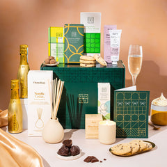 EDEN & CO. Pamper Hamper gift set with a mini Bottega Gold prosecco, dark green gift cylinder, an “Occasion Themed” card, chocolate bars and truffles, a scented candle and Aery hand cream, styled with gold silk against a warm terracotta background.