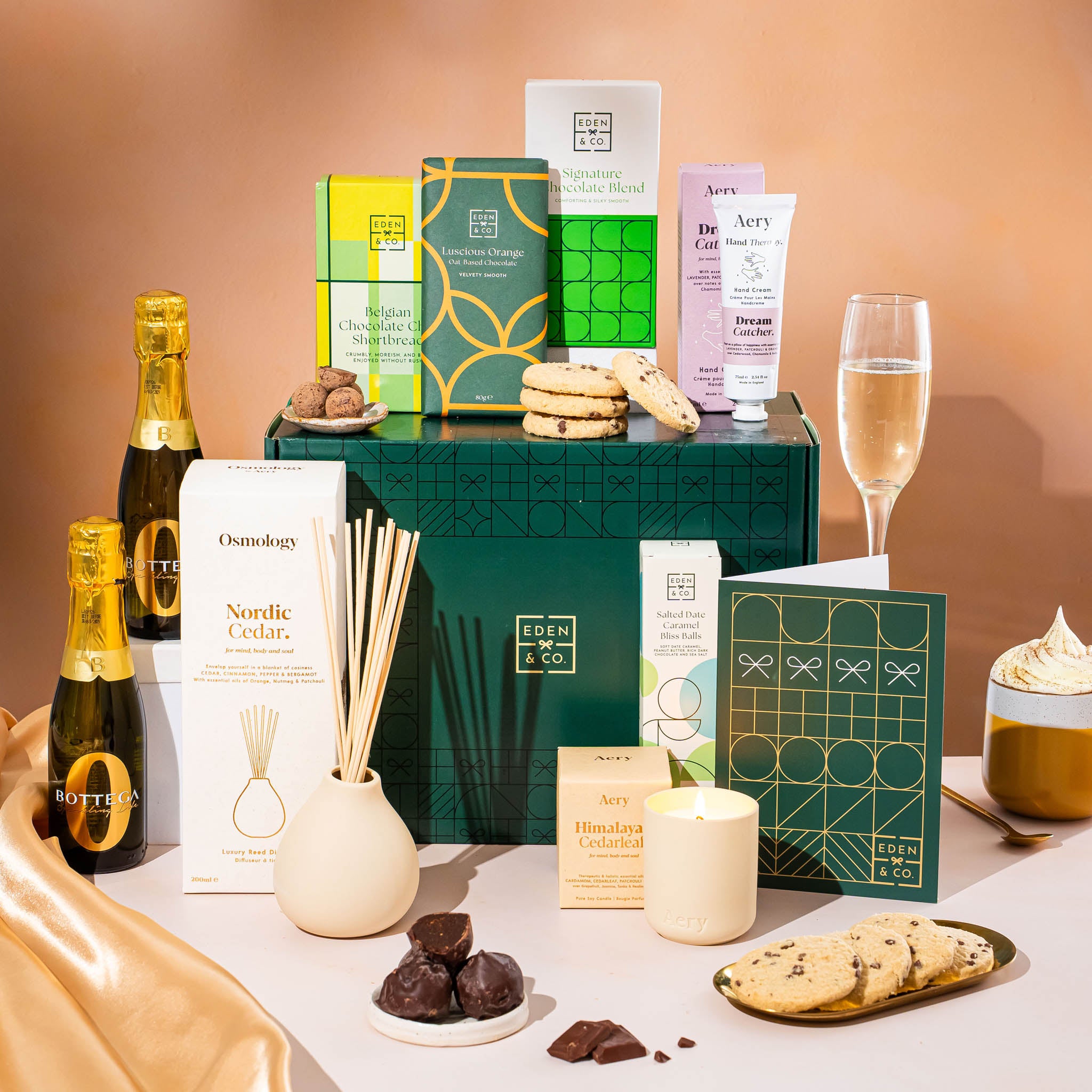 EDEN & CO. Pamper Hamper gift set with a mini Bottega Zero Black prosecco (0% alcohol-free), dark green gift cylinder, an “Occasion Themed” card, chocolate bars and truffles, a scented candle and Aery hand cream, styled with gold silk against a warm terracotta background.