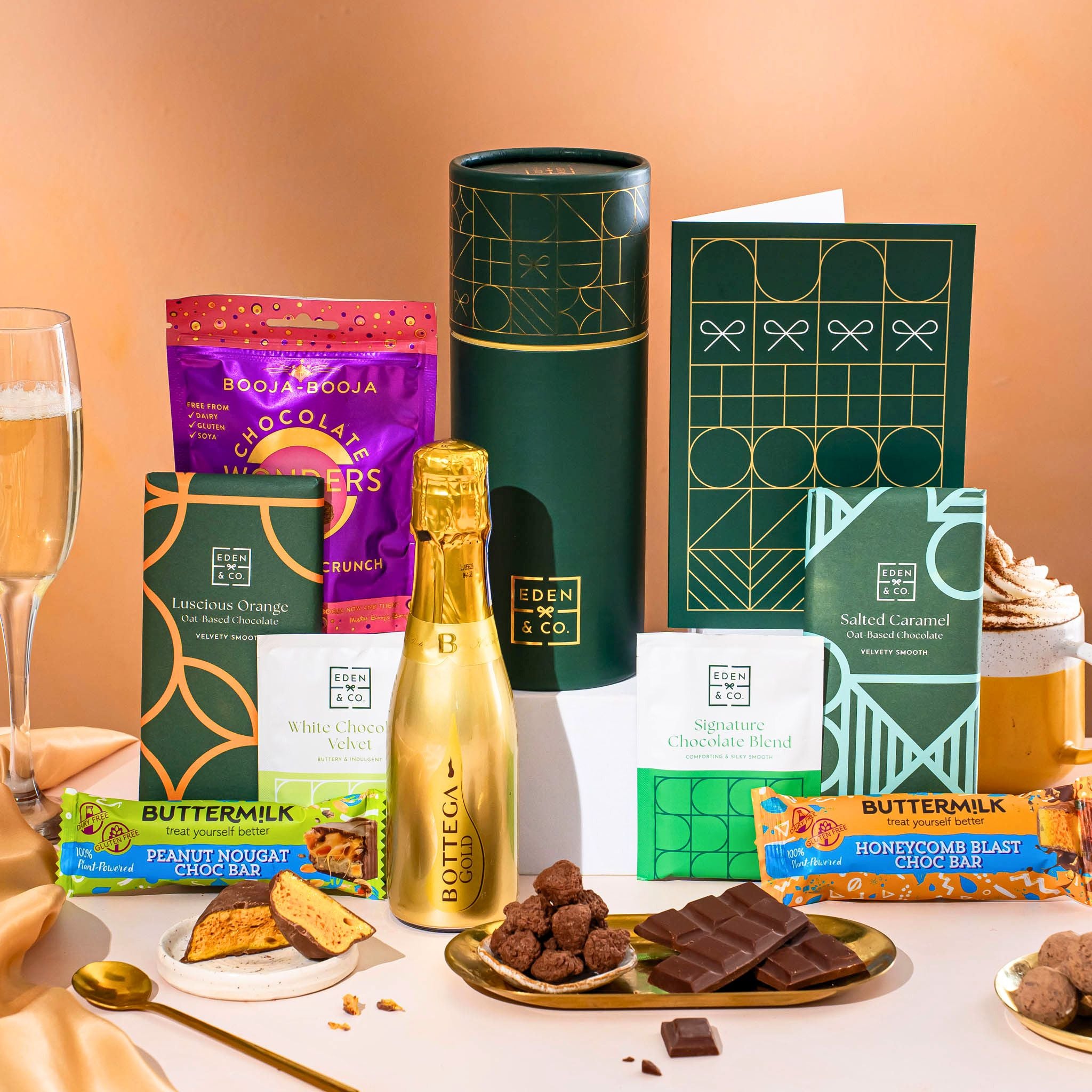 EDEN & CO. plant-based gift hamper with a mini Bottega Gold prosecco, green gift tube, “Occasion Themed” card, chocolate bars in Luscious Orange, White Chocolate Velvet, Signature Chocolate Blend and Salted Caramel, Booja-Booja Chocolate Wonders truffles, Buttermilk Peanut Nougat and Honeycomb Blast bars, chocolate truffles and honeycomb pieces on small plates, and a filled champagne glass, set against a warm terracotta background.