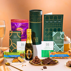 EDEN & CO. plant-based gift hamper with a mini Bottega Zero Black prosecco, green gift tube, “Occasion Themed” card, chocolate bars in Luscious Orange, White Chocolate Velvet, Signature Chocolate Blend and Salted Caramel, Booja-Booja Chocolate Wonders truffles, Buttermilk Peanut Nougat and Honeycomb Blast bars, chocolate truffles and honeycomb pieces on small plates, and a filled champagne glass, set against a warm terracotta background.