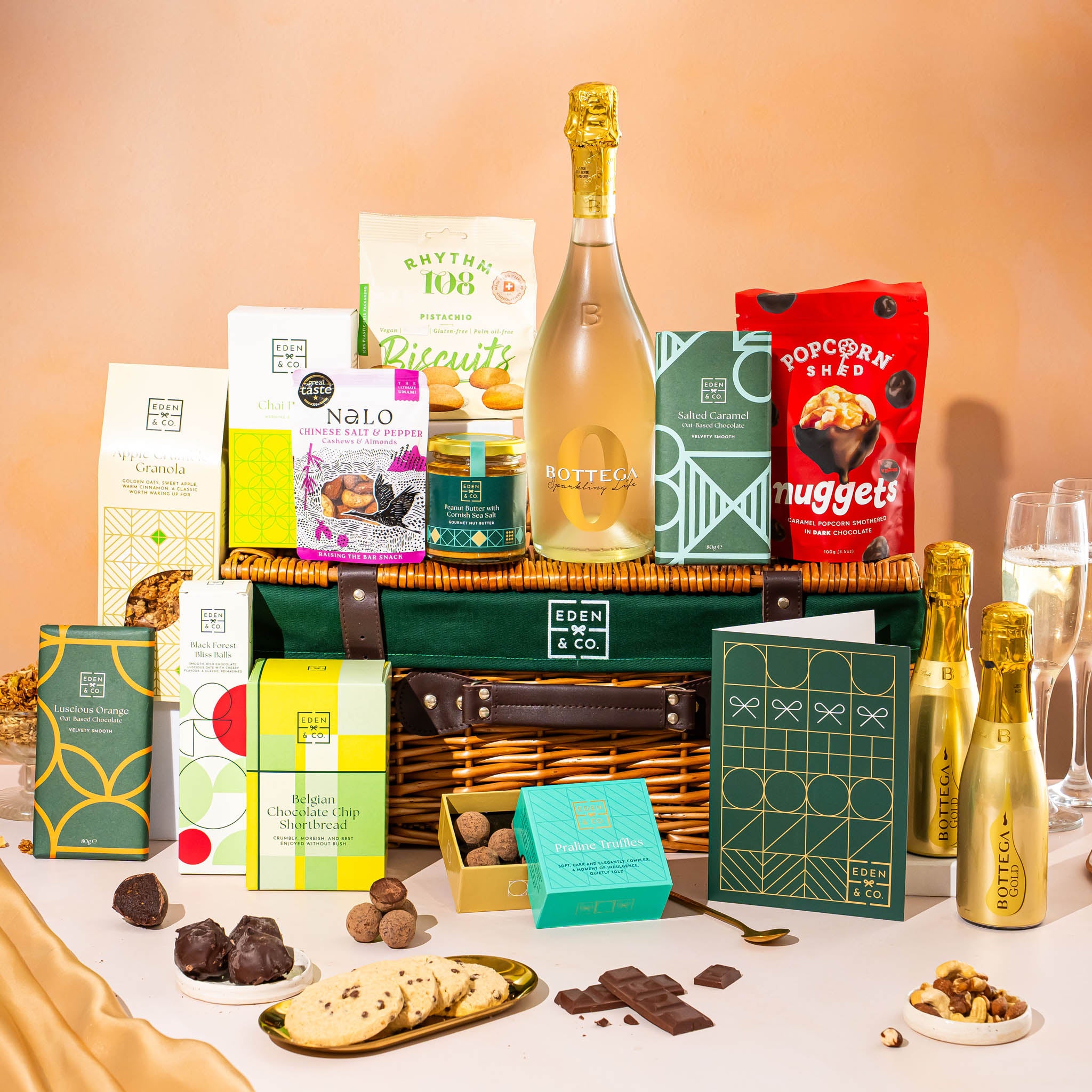 EDEN & CO. Luxury Wicker Hamper filled with a full-size Bottega Gold prosecco, an assortment of chocolate bars, Booja-Booja Chocolate Wonders truffles and Buttermilk snack bars, presented in a dark green trimmed wicker basket with an “Occasion Themed” card, styled against a warm terracotta background.